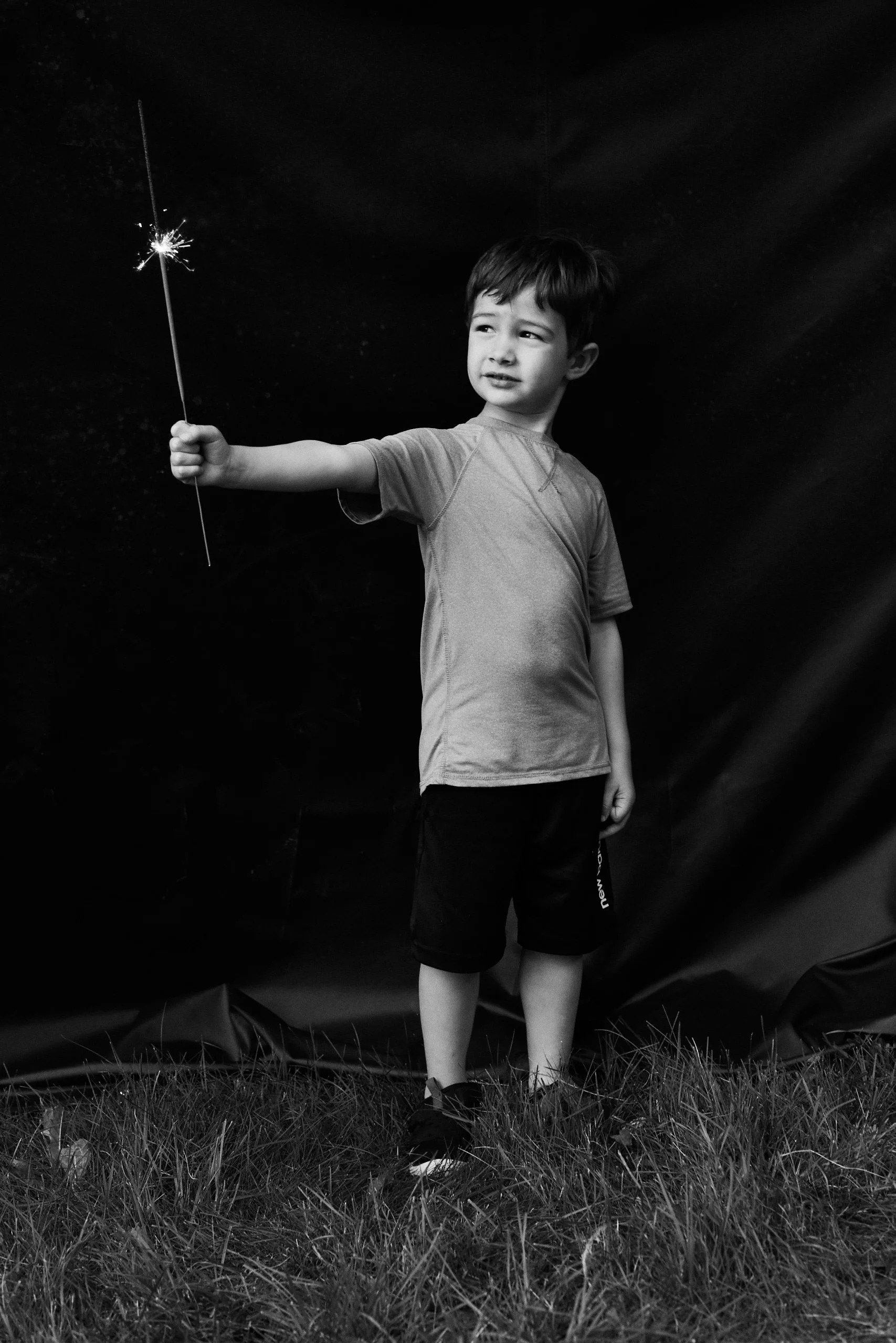 A young boy holding a lit sparkler in a grassy area against a dark sky, looking at the sparkler.