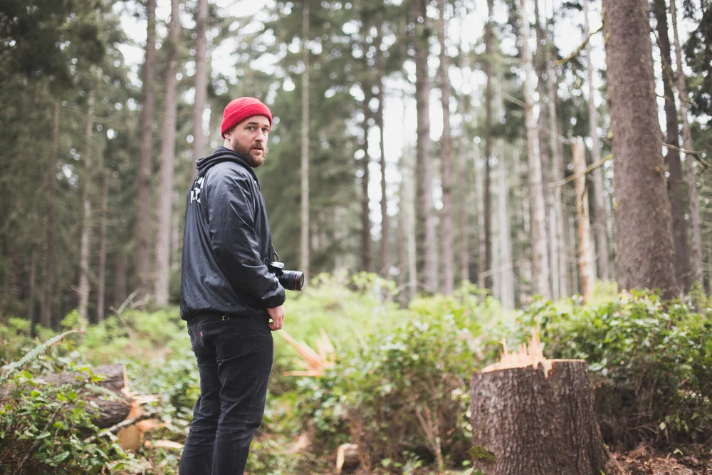 Man with a camera around his neck standing in a forest, looking over his shoulder next to a tree stump.