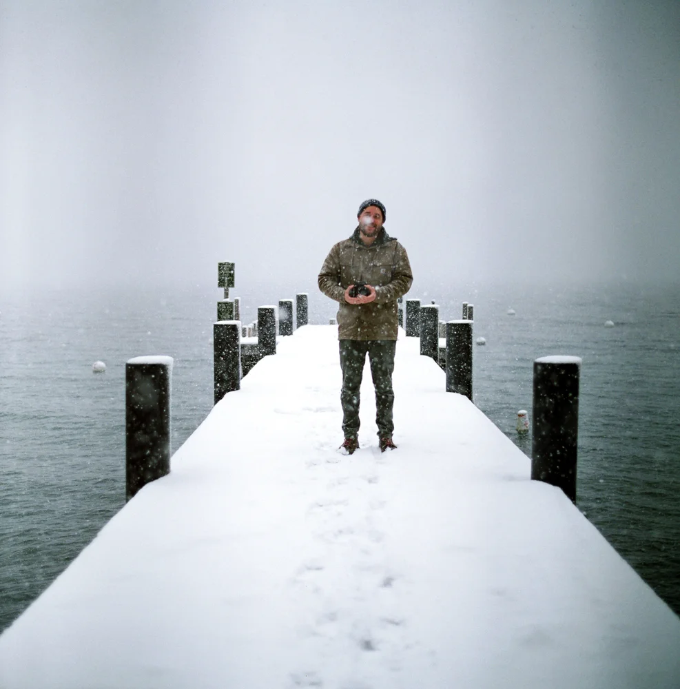 A person standing on a snow-covered dock by a body of water during snowfall, holding a camera.