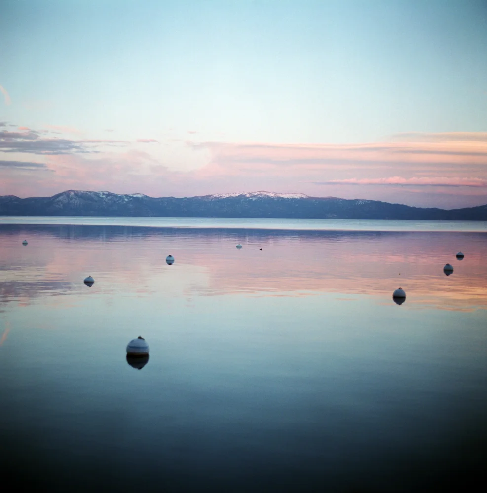 Peaceful scene of a calm lake at sunset or sunrise with distant mountains, pink and blue sky, and scattered buoys floating on the water.