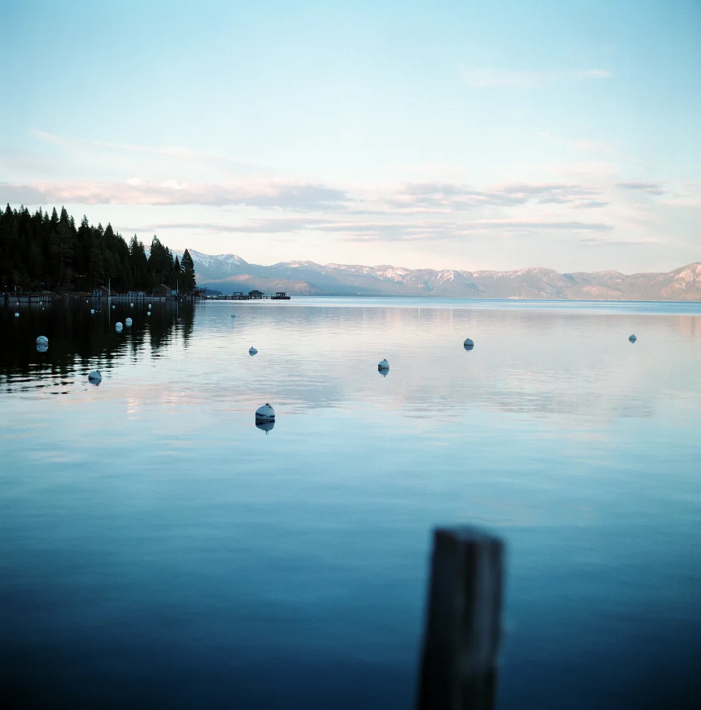 Calm lake with white buoys, forested shoreline on the left, mountain range in the background, and a partly cloudy sky.