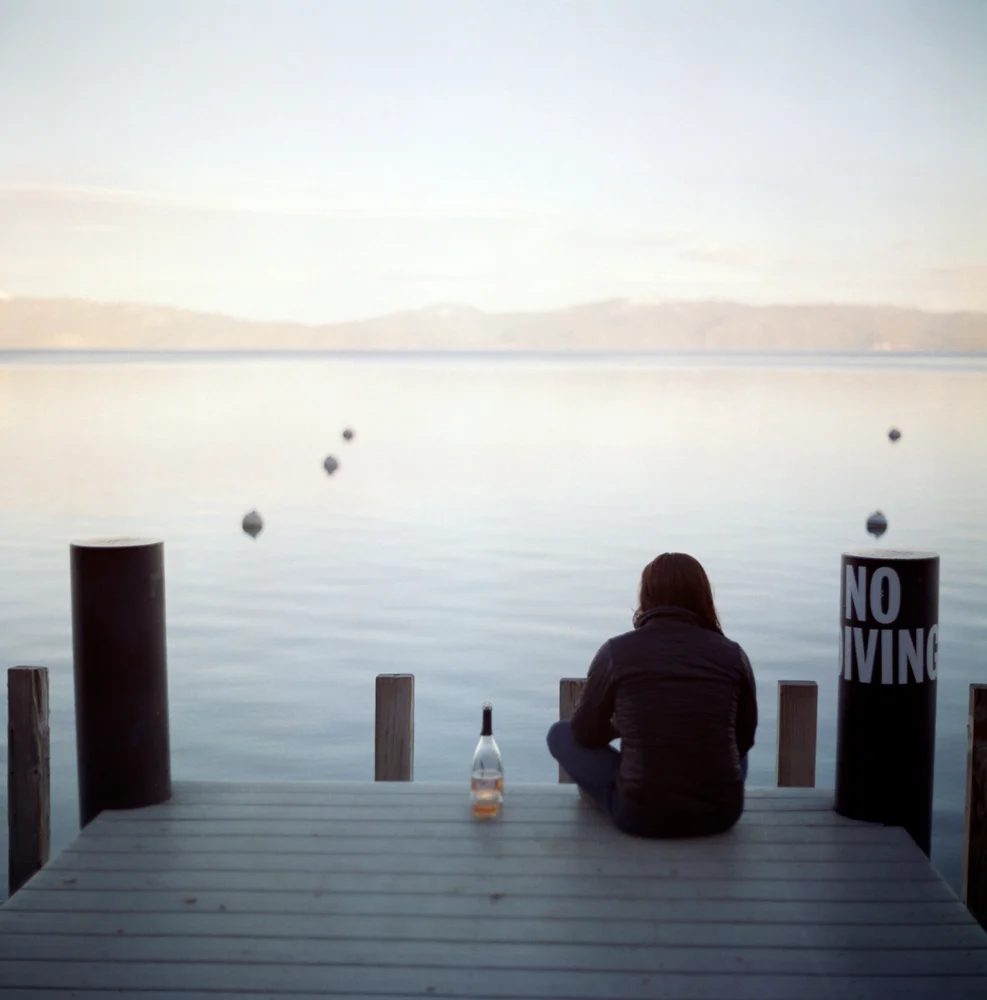A person sitting on a dock near a calm body of water, facing away, with a bottle placed beside them. The scene is peaceful with distant hills and a mostly clear sky. A sign nearby reads 'NO DIVING'.