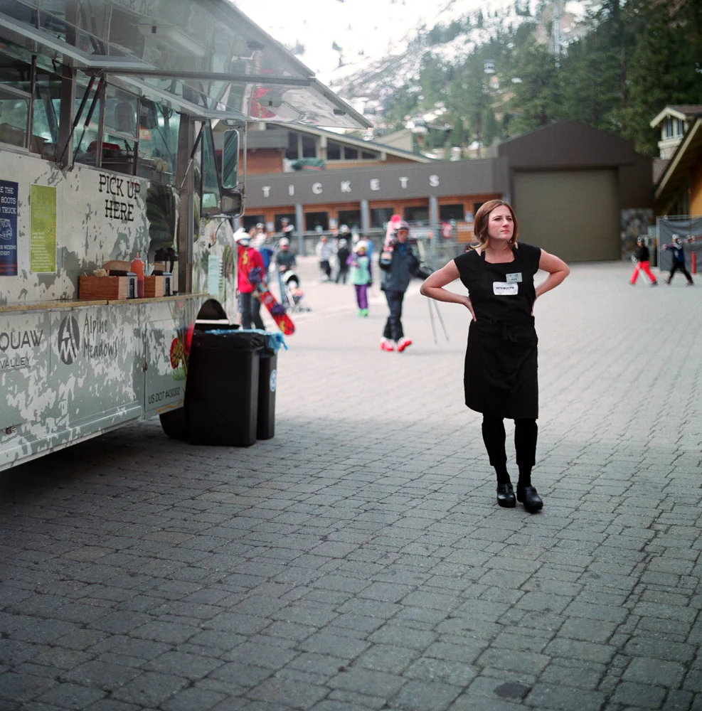 A woman standing with her hands on her hips near a food truck at a ski resort, with a ticket booth and people in winter clothing in the background.