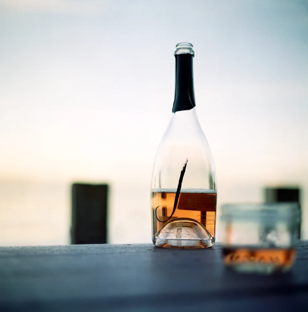 A glass bottle with a black neck and an 'L' on it, filled with a pinkish liquid, on a surface with a small glass of the same liquid next to it, against a blurred background of a sunset or sunrise over water.