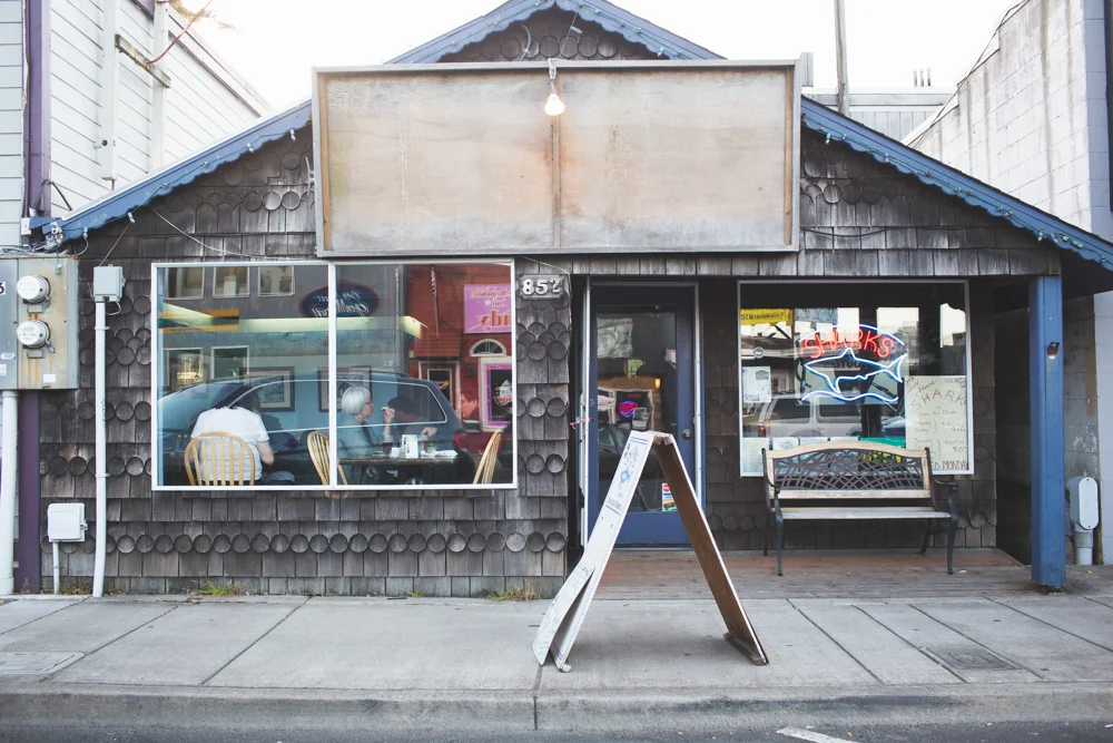 A small restaurant or cafe with a dark wood exterior, large front window showing people inside, a door with a neon shark sign, a bench outside, and a sandwich board sign on the sidewalk.