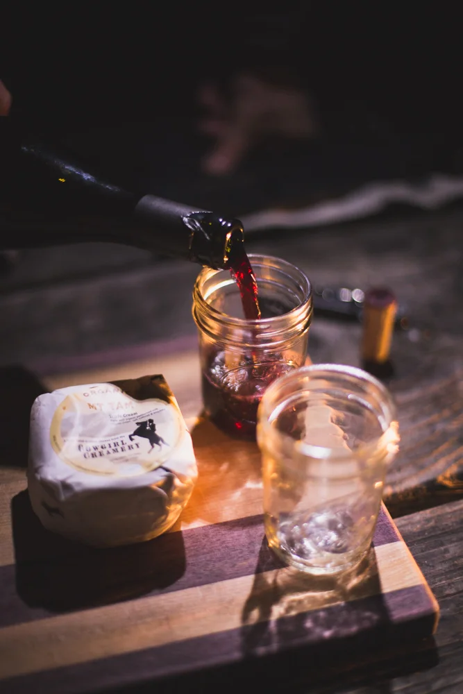 A bottle of red wine being poured into a glass jar with two empty glasses on a wooden cutting board, with a round cheese on the side, all placed on a wooden surface.