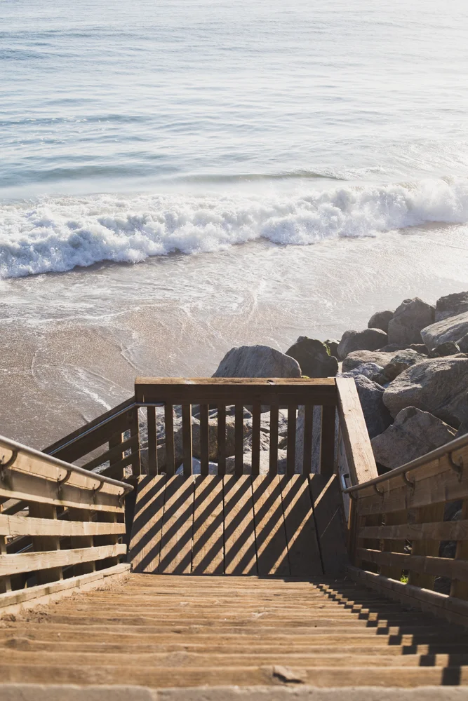 Wooden staircase leading down to a beach shoreline with rocks and gentle waves, overlooking the ocean.