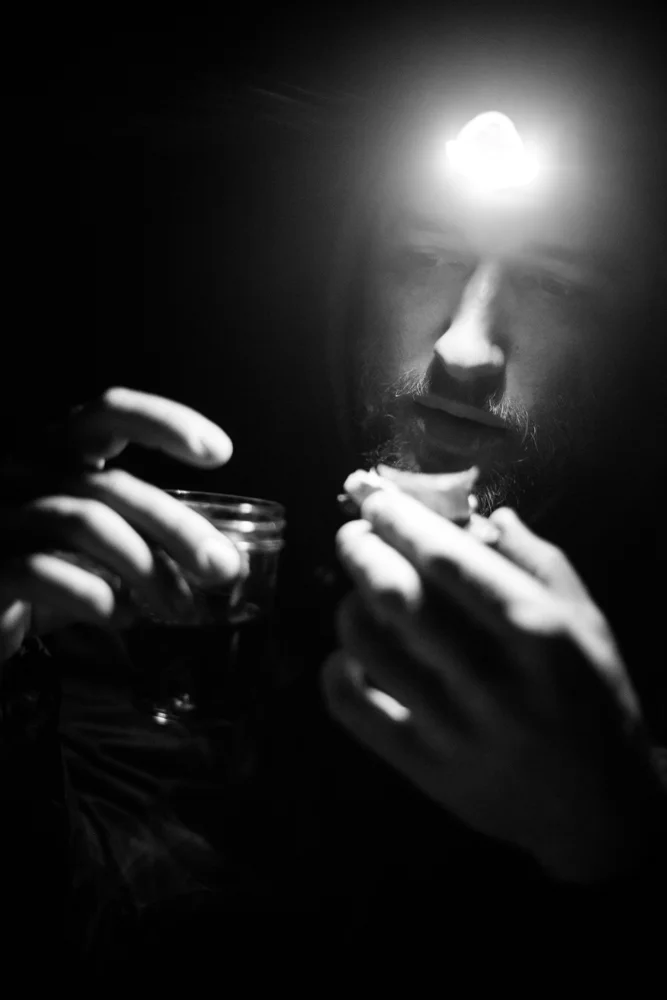 Black and white photo of a man with a headlamp, holding a small jar and a spoon, eating something in a dark setting.