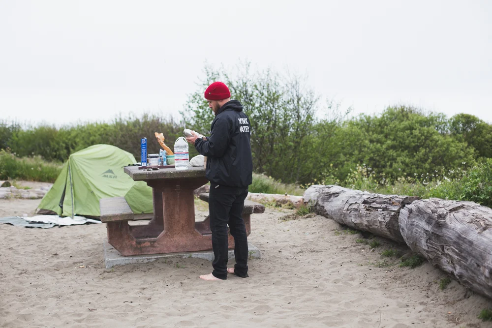 Man in black hoodie and red beanie standing barefoot by a picnic table on a sandy area, with a green tent and a large fallen log in the background.