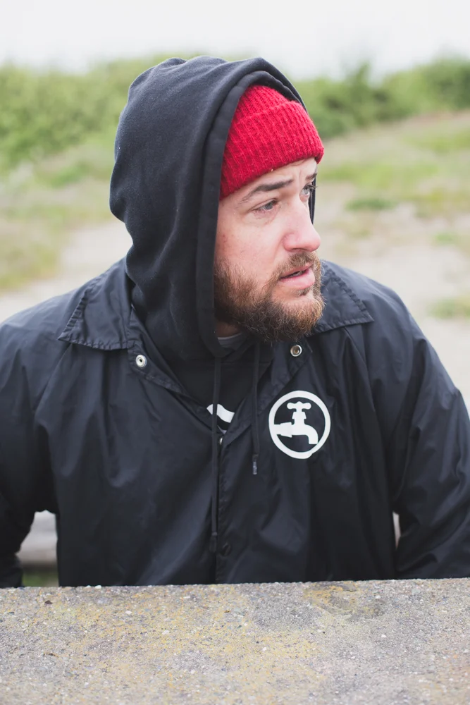 A man with a beard, wearing a red beanie, a black hooded jacket with a faucet logo, and a black hoodie, looking to the side outdoors.