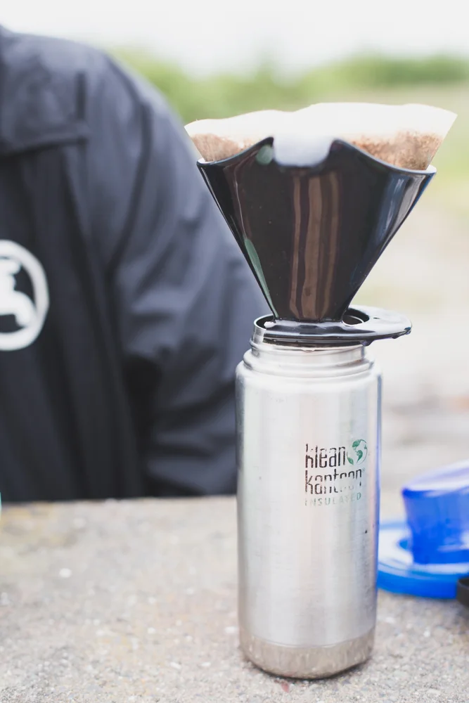 Coffee being poured through a pour-over coffee maker into a metal insulated bottle outdoors.