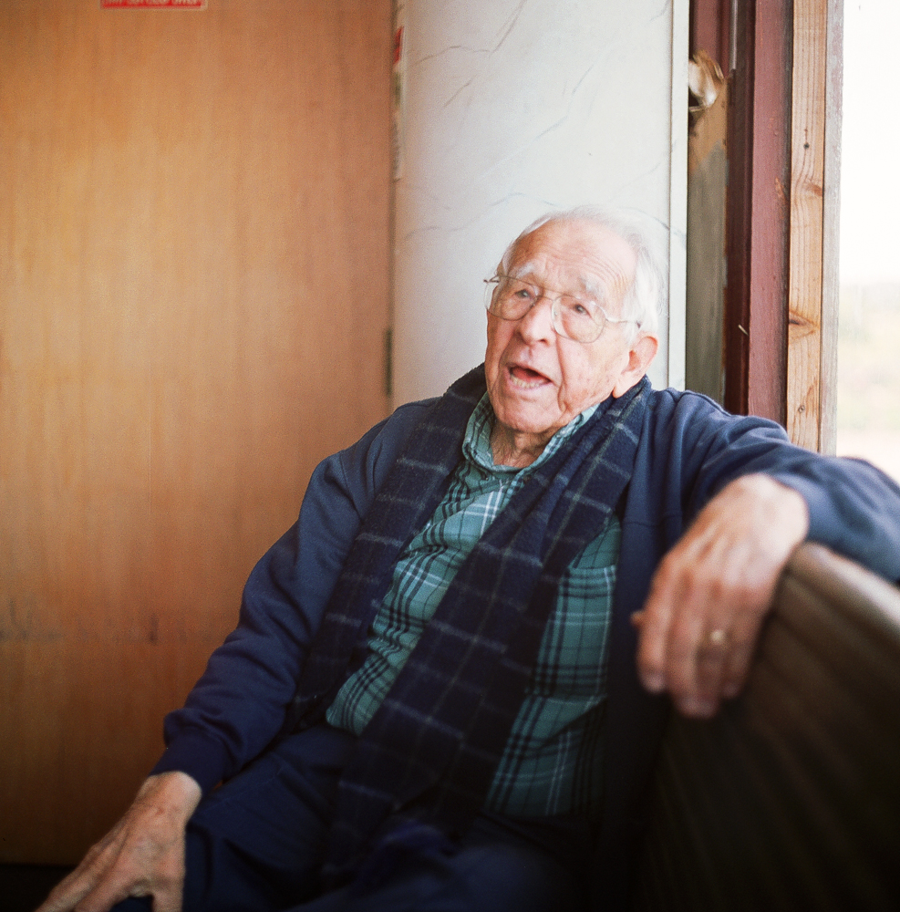 An elderly man with glasses, wearing a plaid shirt and a dark scarf, is sitting on a bench near a window, talking or singing.