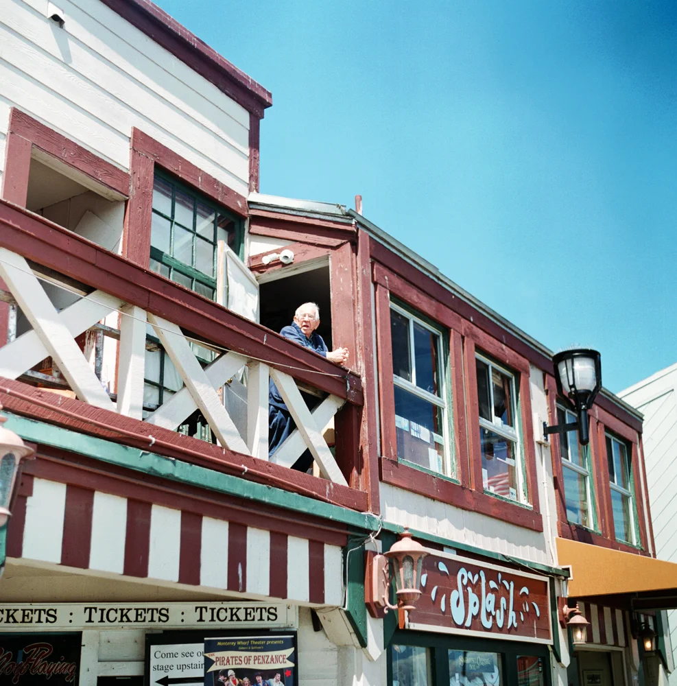 Older man standing on a balcony of a two-story building with striped and solid colored exterior, looking out. The building has large windows and a sign that reads 'Splash' in stylized letters. Clear blue sky in the background.
