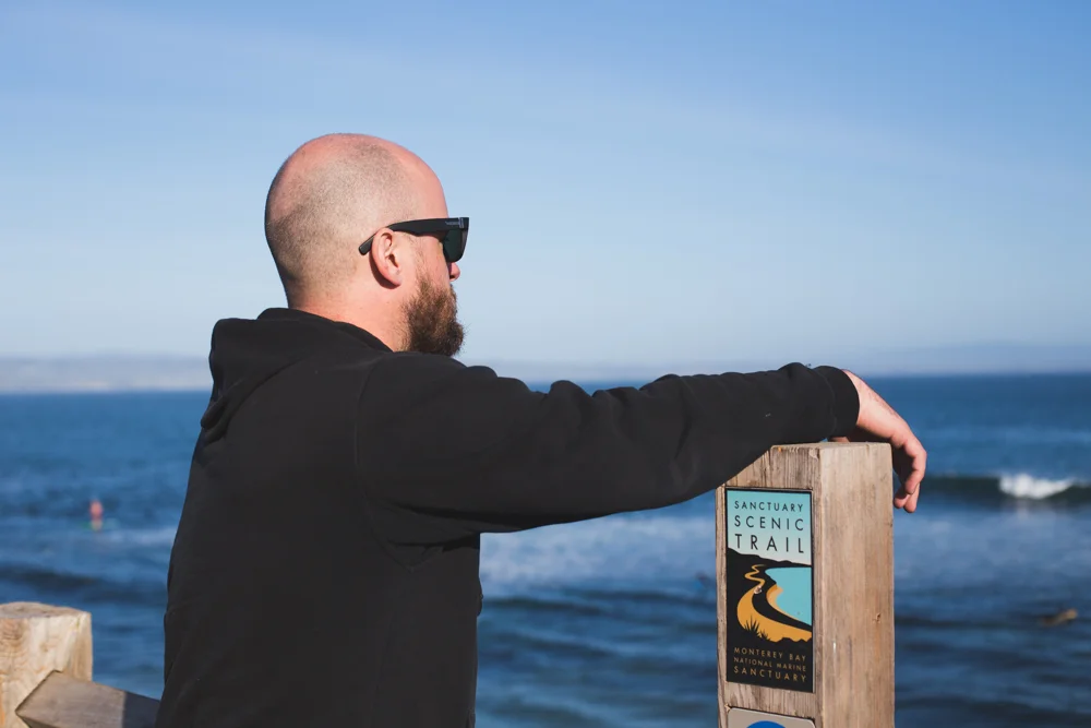 Man with sunglasses and beard leaning on a wooden sign marked 'Sanctuary Scenic Trail' overlooking the ocean.