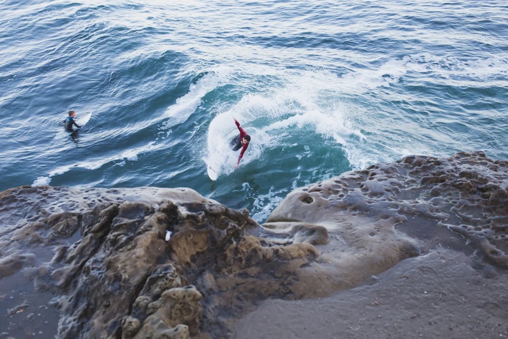 Surfer riding a wave near rocky shoreline, with another person in the water