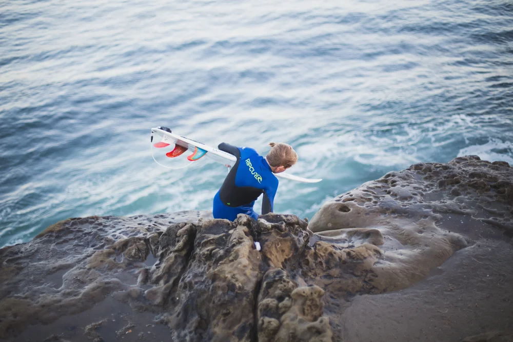 Person in a wetsuit climbing over rocks near the water, holding a surfboard.