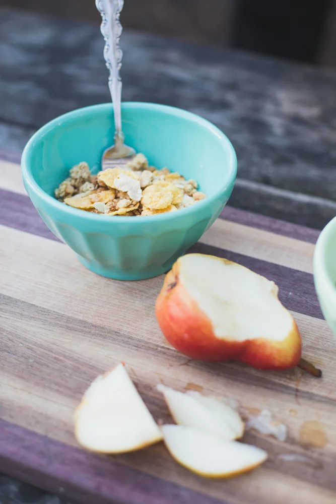 A bowl of cereal with milk being poured into it, placed on a wooden surface next to a partially eaten apple with slices, also on the wooden surface.