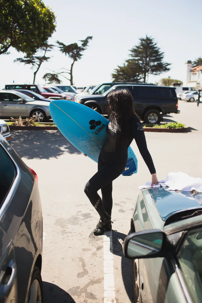 A woman in a black wetsuit carrying a blue surfboard walks in a parking lot.