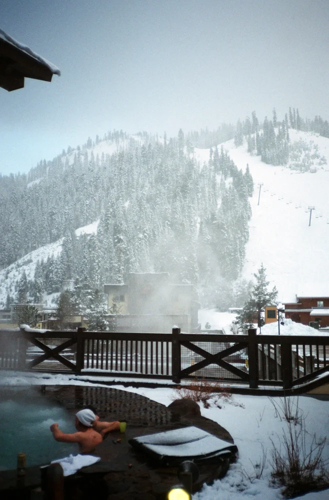 A snowy mountain landscape with ski slopes, a building, and trees in the background. In the foreground, a man is relaxing in an outdoor hot tub, wearing swimming goggles and an arm float, with snow around the area.