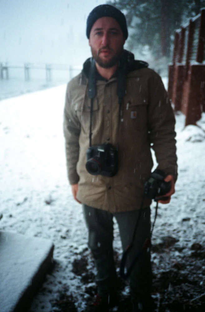 A man standing outdoors in snowy weather, wearing a brown jacket, black beanie, and holding a camera with a strap around his neck.