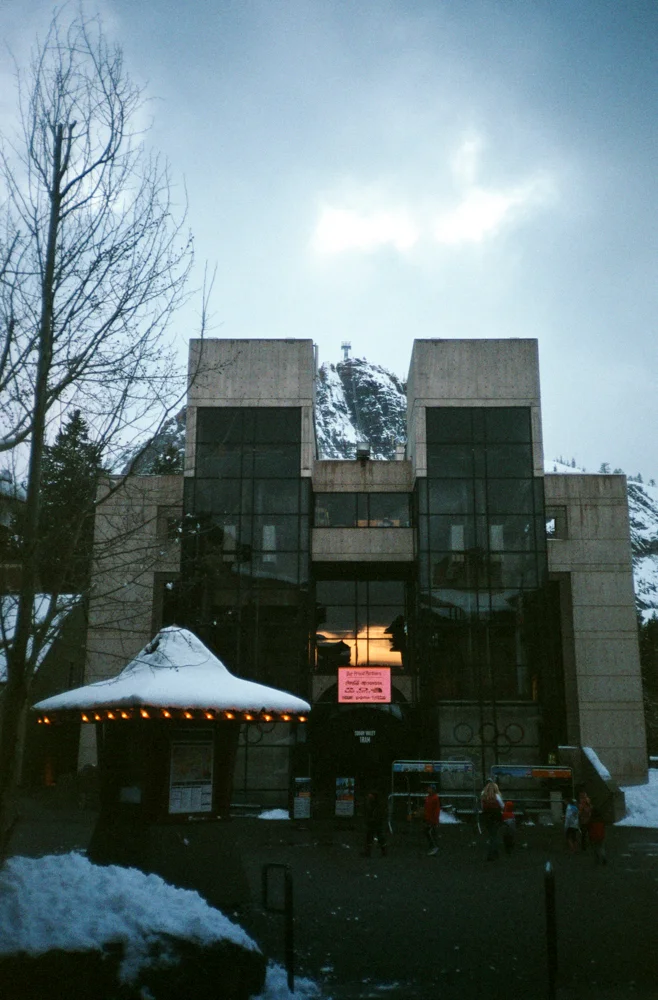 Modern building with large glass windows, snow on the ground, and a mountain in the background at sunset. Several people walking nearby.