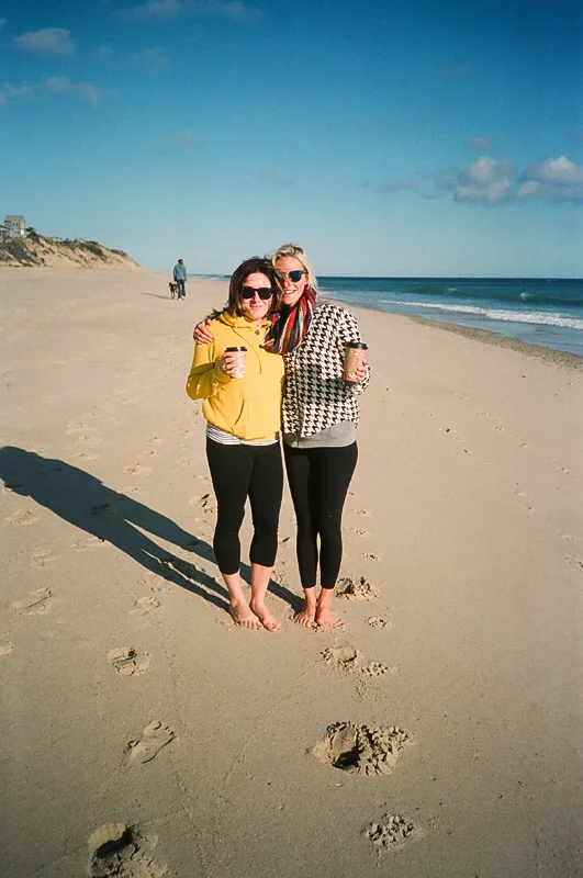 Two women standing on a sandy beach, smiling, with ocean waves and a clear blue sky in the background, holding drinks.