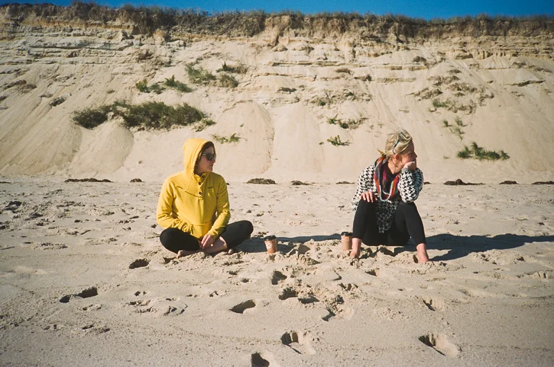 Two women sitting on the sandy beach near cliffs, one wearing a yellow hoodie and sunglasses, the other in black pants and a patterned sweater, with cups and a small sandcastle between them.