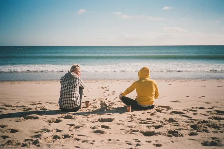 Two people sitting on the sandy beach facing the ocean, with small cups beside them, under a partly cloudy sky.