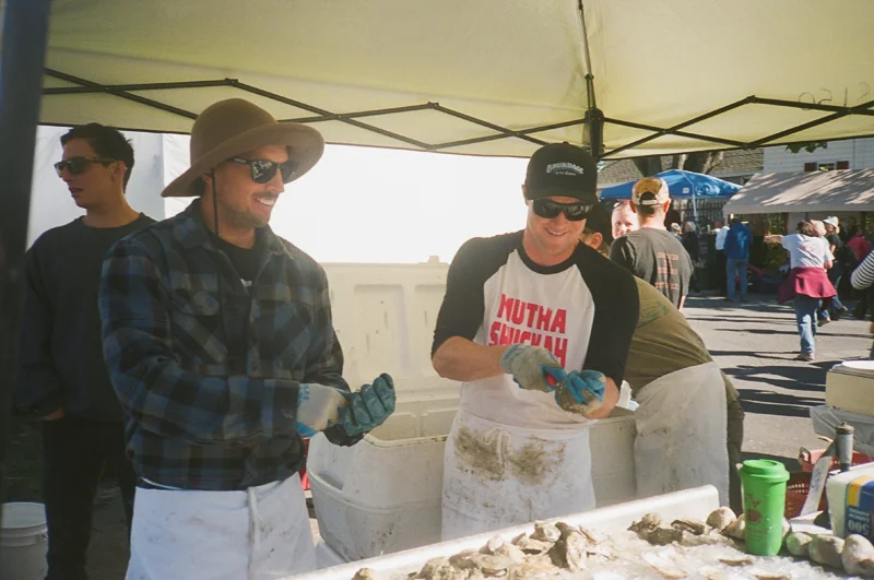 Two men at an outdoor seafood market under a tent, wearing gloves and aprons, handling seafood. One man is wearing a wide-brimmed hat and sunglasses, while the other is in a baseball cap.