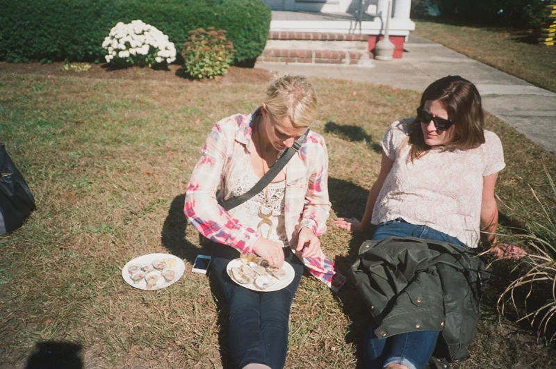 Two women are sitting on the grass in front of a house, with one woman opening a shellfish shell on a plate, and the other woman watching. There is a plate of shells on the ground nearby.