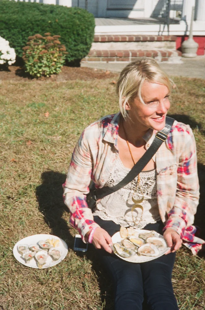 Woman sitting outdoors on grass, holding a plate of oysters, with an additional plate of oysters nearby, smiling in sunlight.