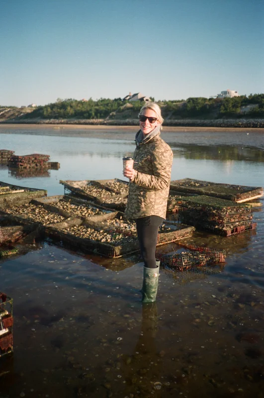 Woman standing in shallow water at the beach, holding a coffee cup, wearing sunglasses, camouflage jacket, and rain boots, with oyster beds and a distant shoreline with houses and green trees under a clear blue sky.