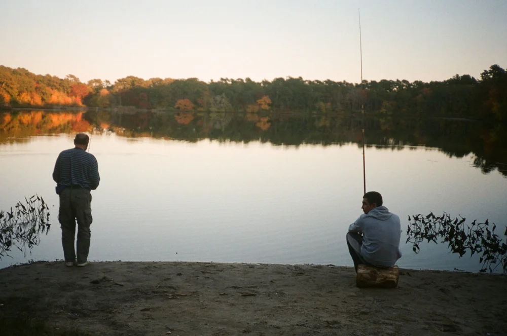 Two men fishing by a calm lake with trees in the background, one standing and one sitting on a log, during sunset.