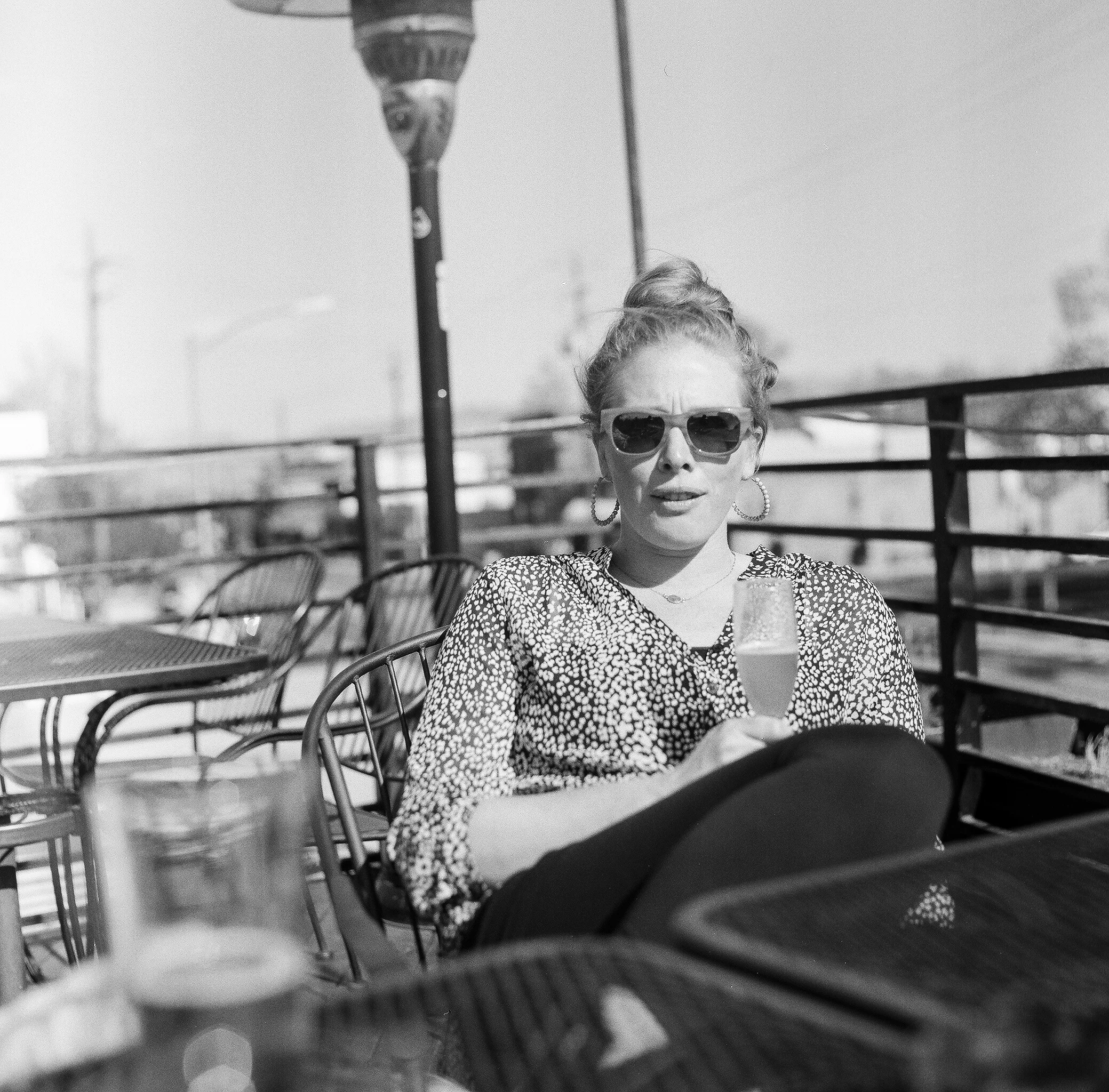 A woman with her hair in a bun, wearing sunglasses, hoop earrings, and a patterned top, is sitting outside at a table. She is holding a glass of a bubbly drink and appears to be speaking or reacting. There are chairs, tables, and a railing in the bac