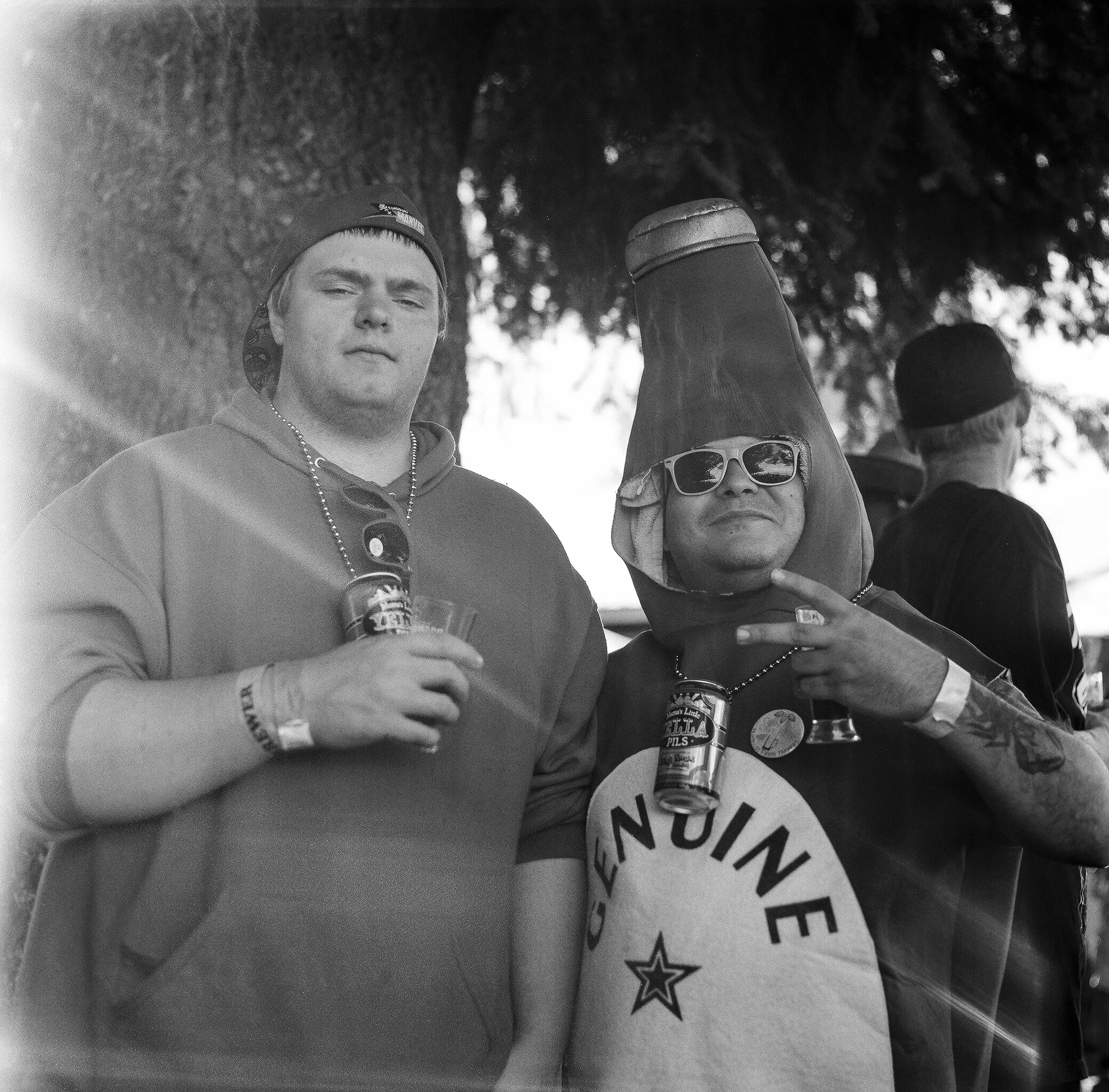 Two men at an outdoor event, one dressed in casual clothing with a hoodie and beanie, the other in a costume featuring a large hat, sunglasses, and a shirt with a star and the word "Genuine." They are holding drinks and making peace signs, with trees