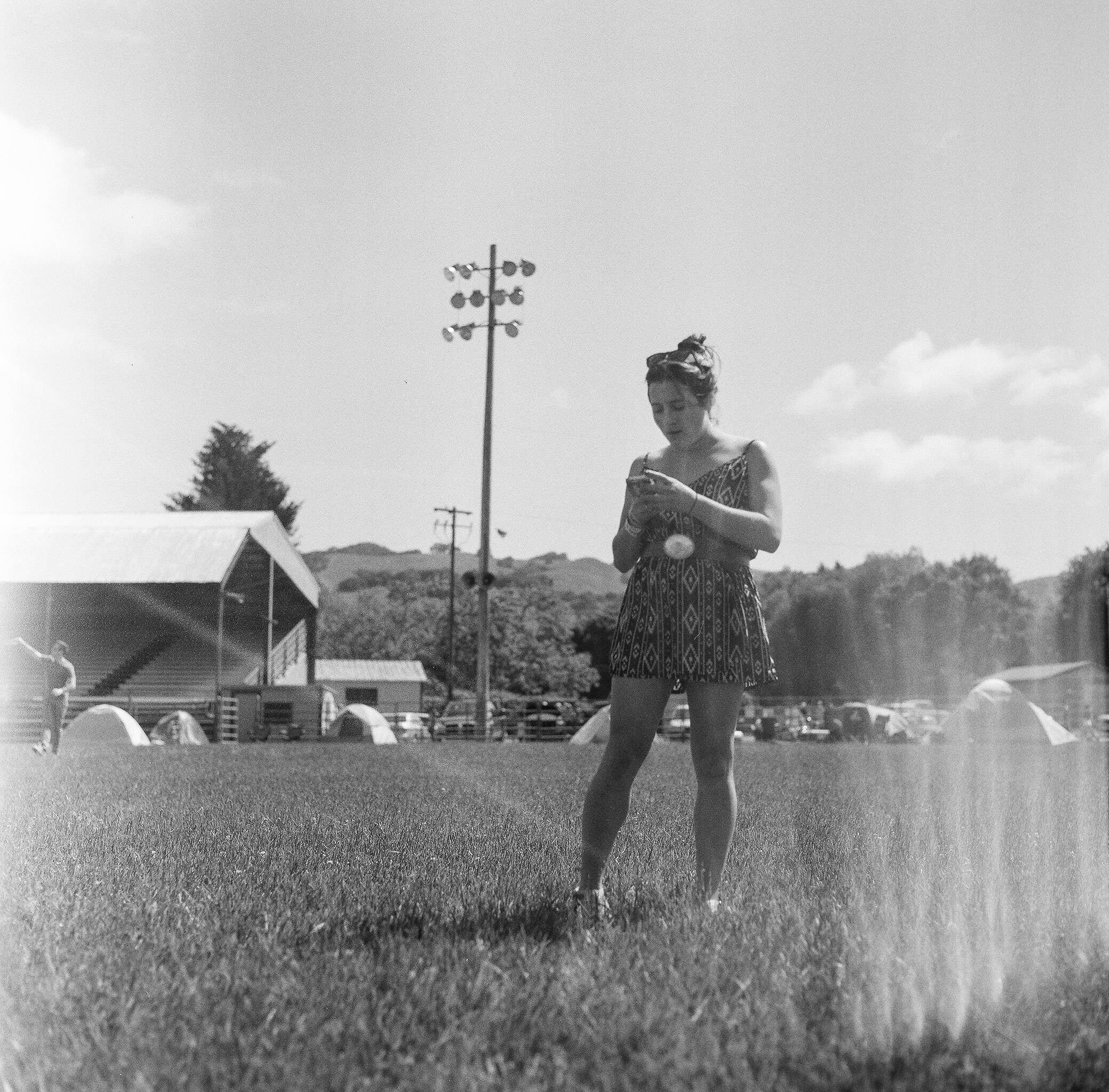 A woman in a patterned dress standing on a grassy field, looking at her phone, with tents, a building, a power pole, and hills in the background on a sunny day.