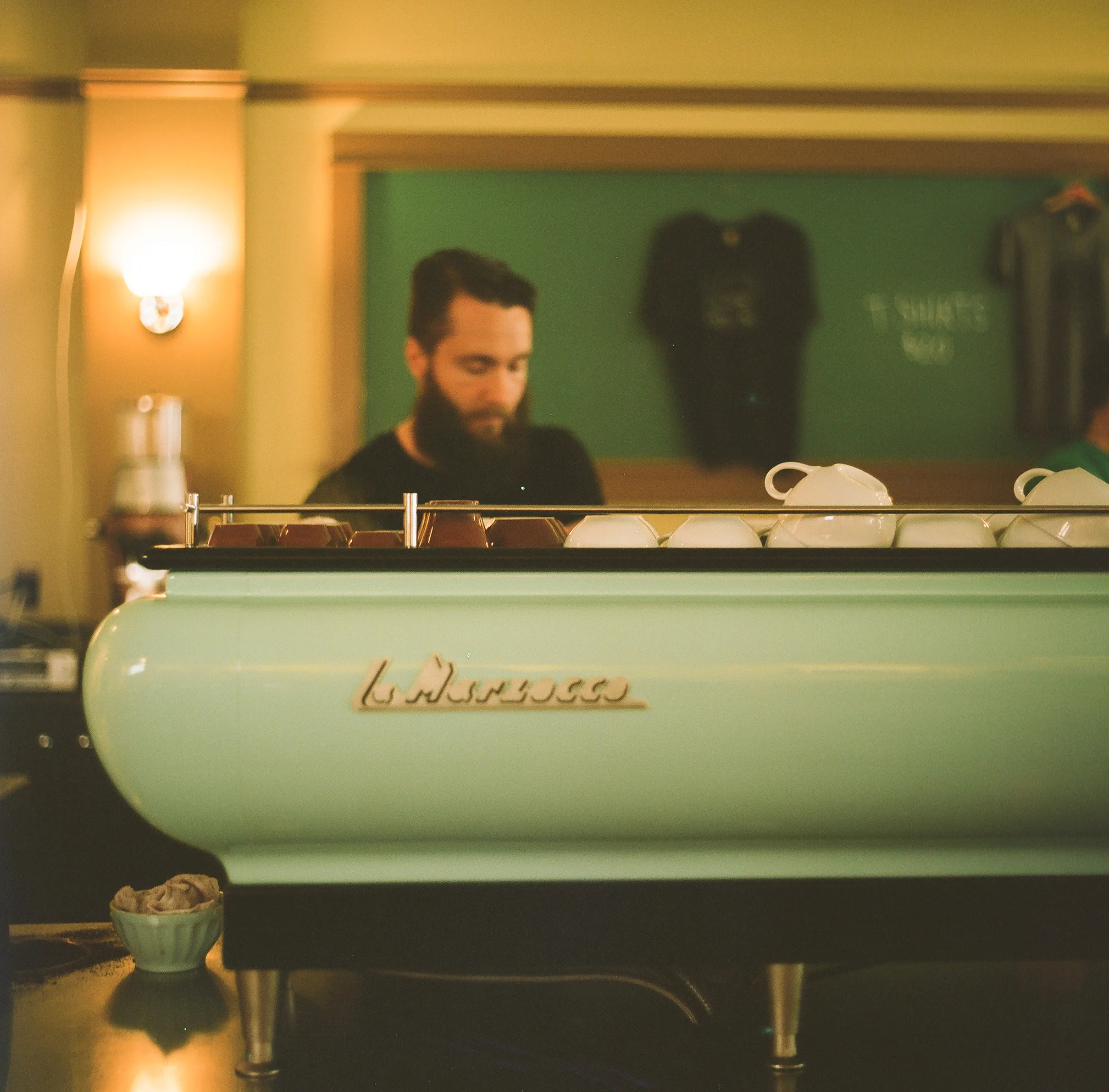 A barista with a beard making coffee behind a vintage mint-green La Marzocco espresso machine, in a cozy coffee shop.