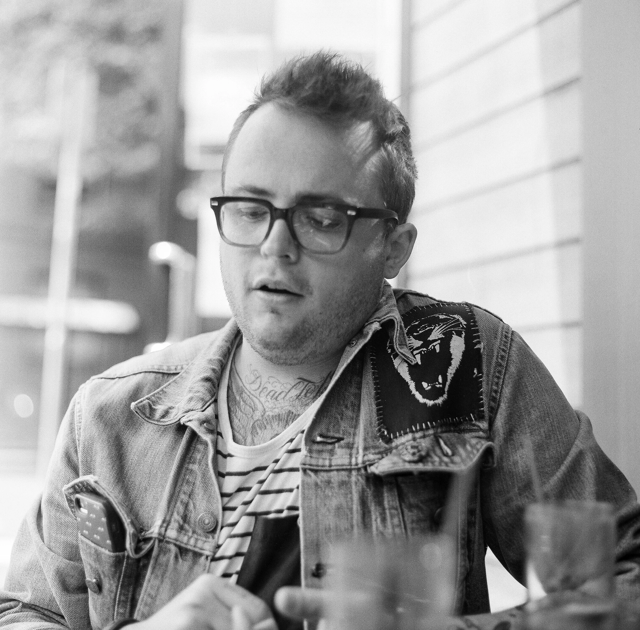 Black-and-white photo of a young man with glasses, tattoos on his neck, wearing a denim jacket with an embroidered patch, looking down at his phone, sitting at a table in an outdoor setting.