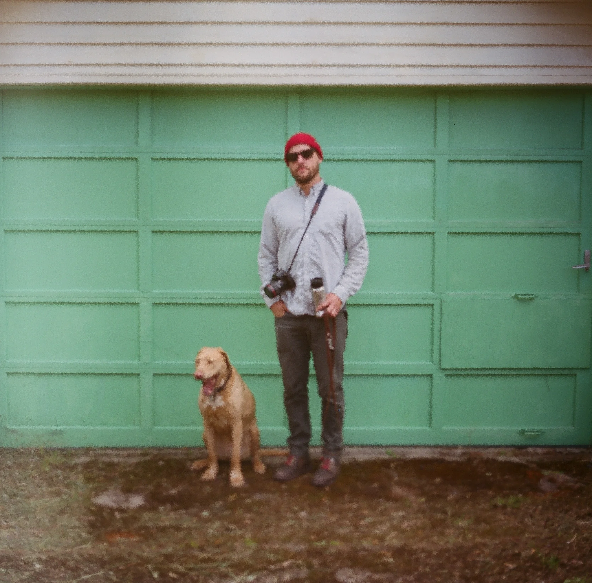 Man standing with a golden retriever dog in front of a green garage door, holding a water bottle, with a camera around his neck and sunglasses, wearing a red beanie and casual clothing.