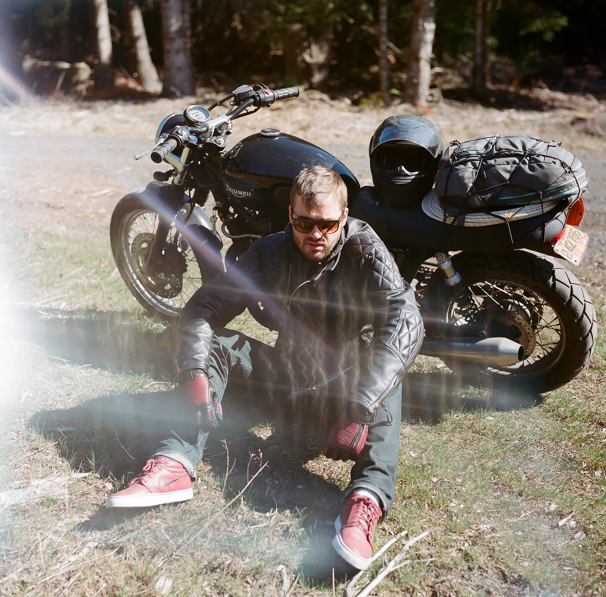 A man sitting on the grass next to a black Triumph motorcycle with a helmet and backpack strapped to back, in a wooded outdoor setting.