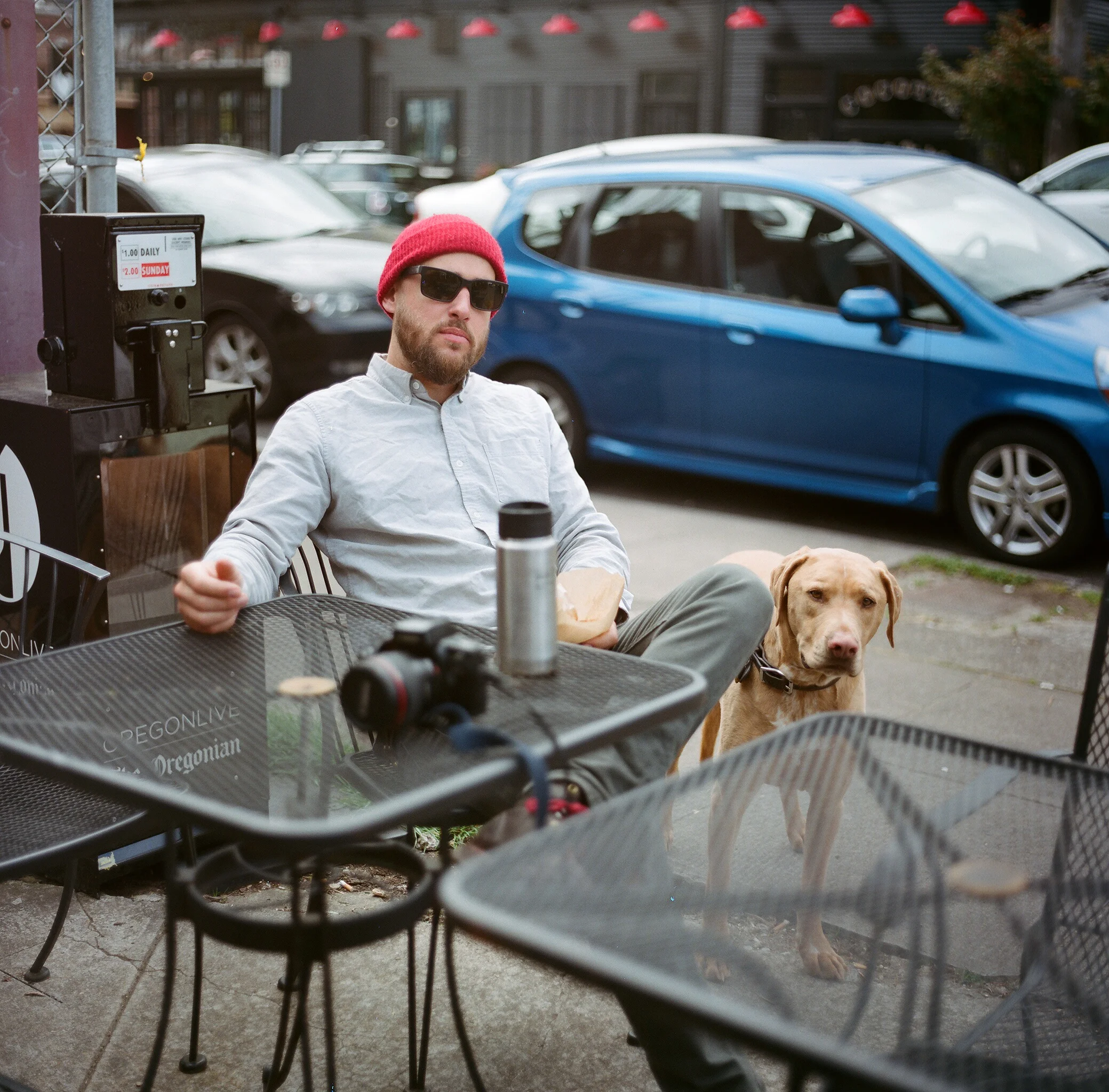 A man with a red beanie, sunglasses, and a light gray shirt sitting at an outdoor cafe table with a Labrador Retriever dog standing next to him.