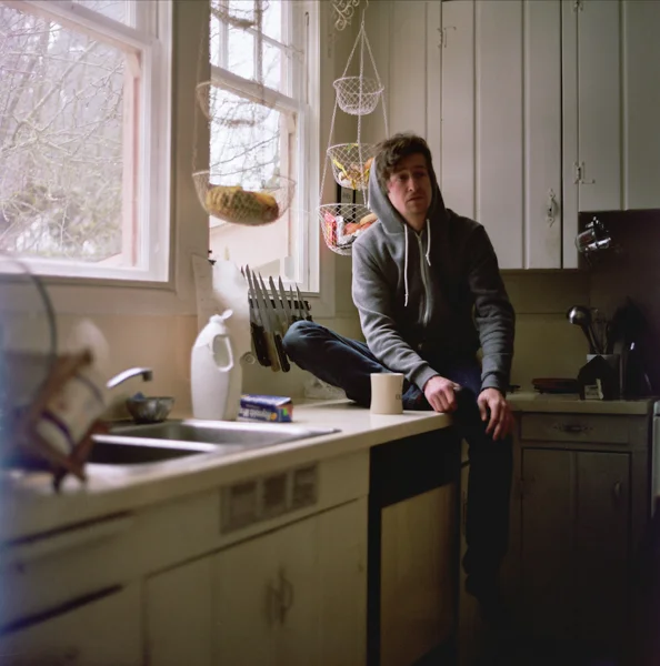 A young man with dark hair wearing a gray hoodie sitting on a kitchen counter next to a window in a rustic kitchen with white cabinets and hanging baskets, holding a mug.