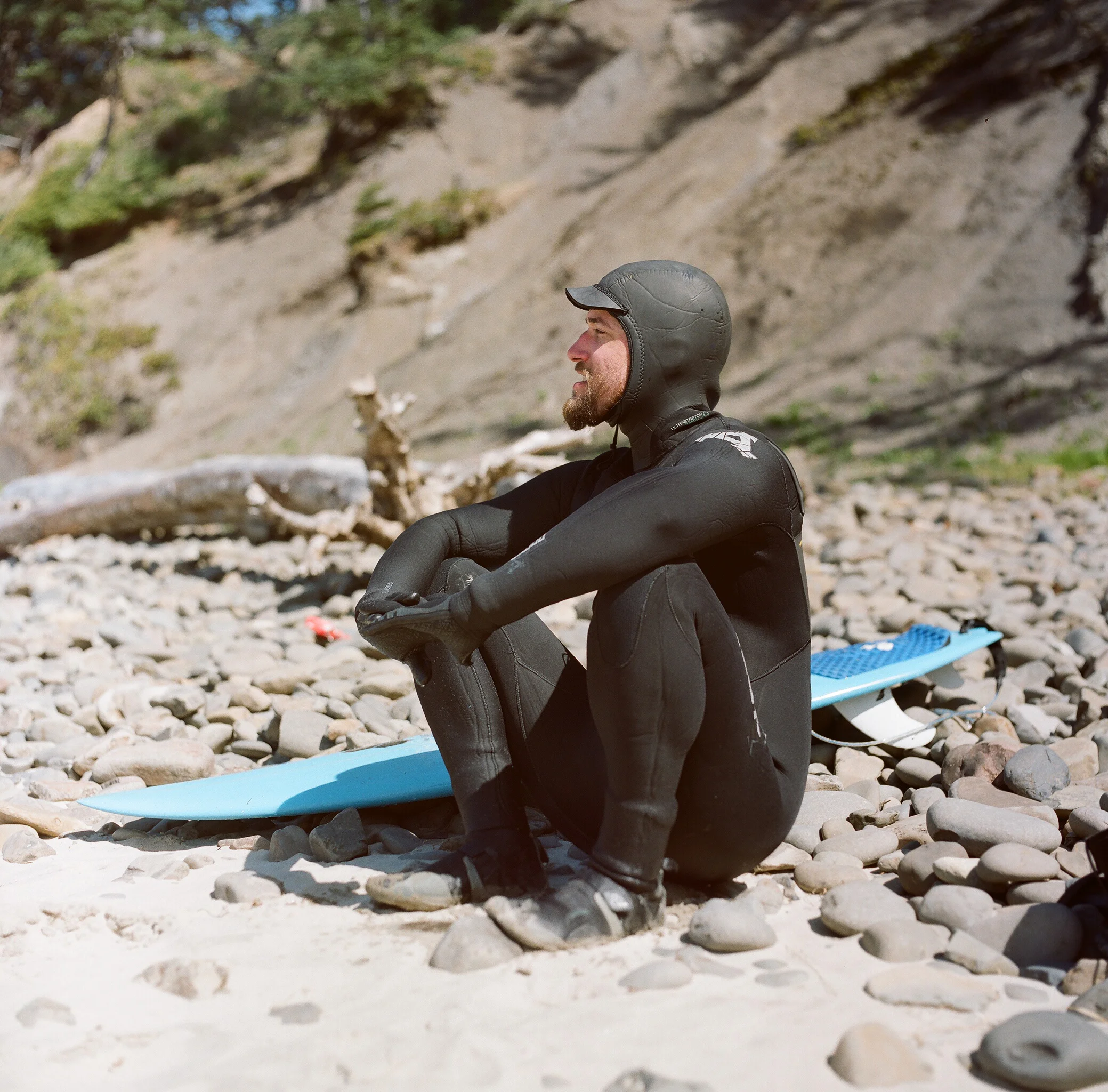 A man dressed in a black wetsuit and helmet sits on a rocky beach with a blue surfboard beside him, looking out towards the water.