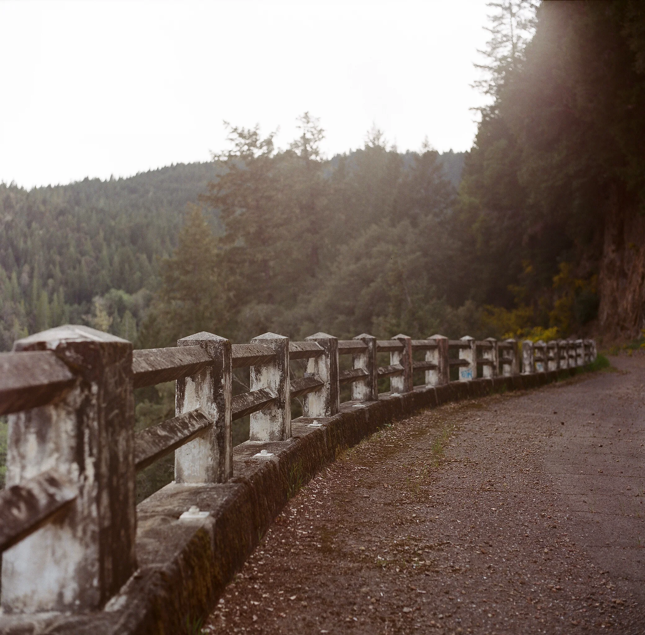 A winding mountain road with a weathered concrete guardrail, surrounded by dense forest and distant hills, under a cloudy sky.