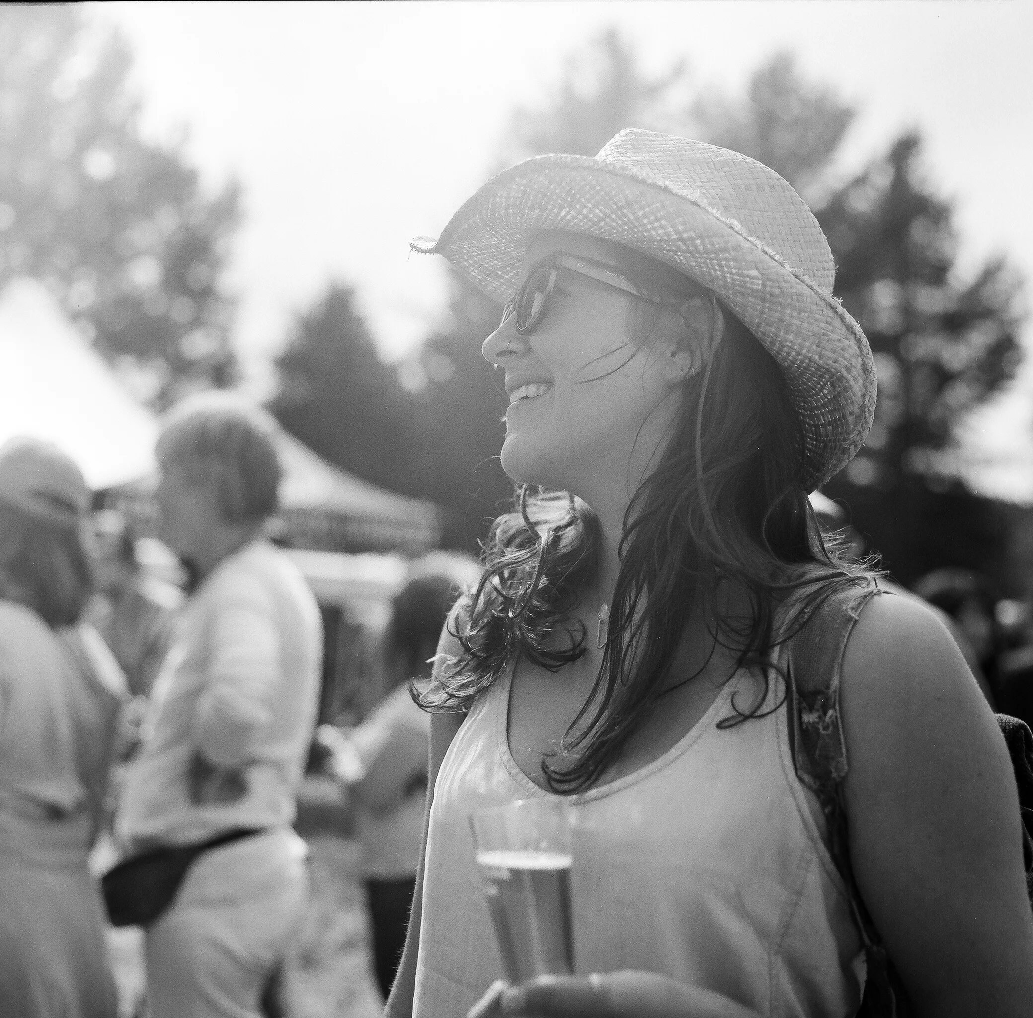 A woman with long hair wearing sunglasses, a straw hat, and a sleeveless top, holding a glass of beer, at an outdoor event.