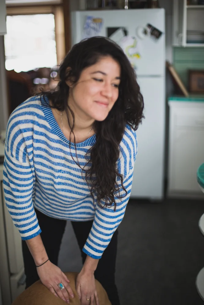 Woman with long dark hair in a blue and white striped sweater leaning over a brown dog in a kitchen.