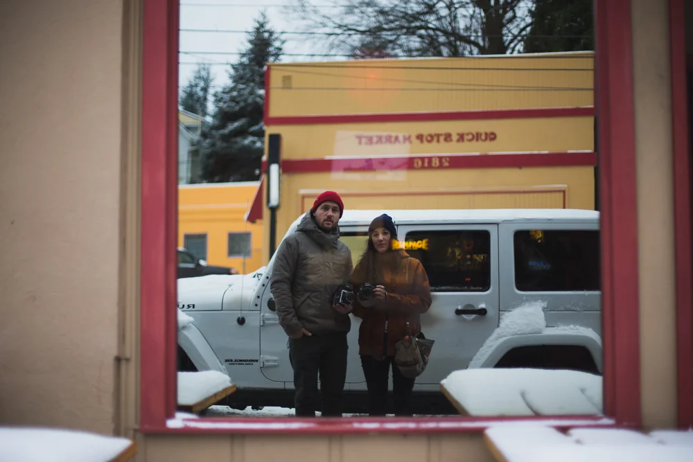 Two people are taking a photo of themselves in a mirror, with a snowy street, a white Jeep, and colorful buildings in the background.