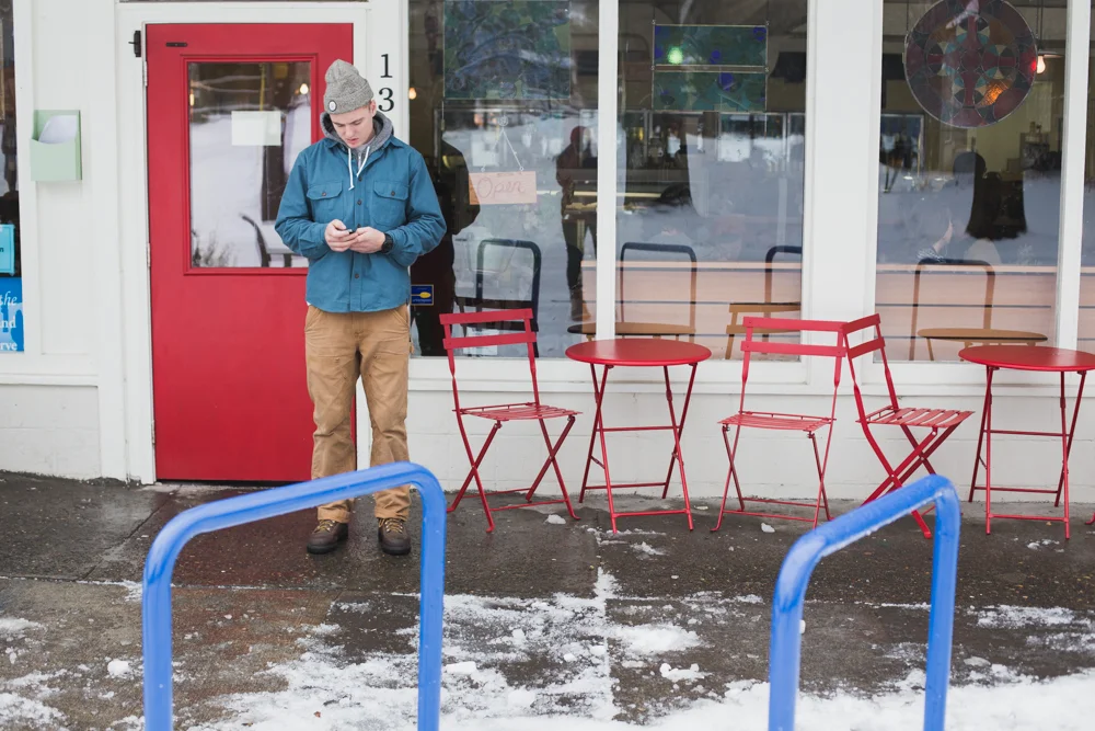 A young man in a gray beanie, blue jacket, and tan pants stands outside a café, looking at his phone. The café has a red door, large windows, and red metal chairs and tables outside. The ground is wet with patches of snow.