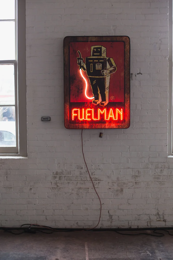 Neon sign featuring a cartoon robot holding a wire, with the word 'FUELMAN' in bright red neon lights, mounted on a white brick wall next to windows.