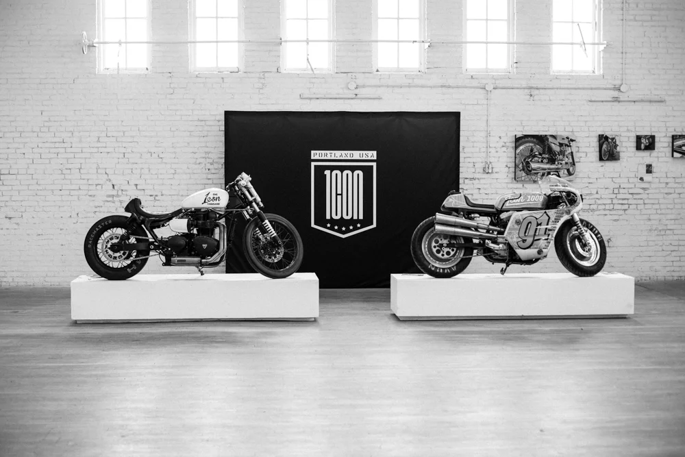 Two vintage motorcycles on display pedestals in an art gallery with a brick wall and artwork in the background. A logo reading 'Portland USA 100M' is on a black banner behind the motorcycles.